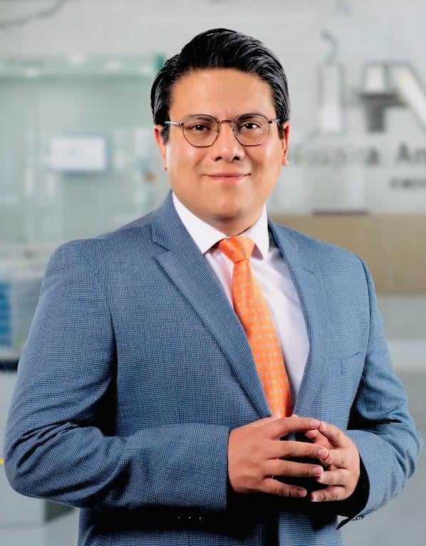 Professional headshot of a man wearing a blue blazer, light pink shirt, and orange tie, standing in a modern office setting